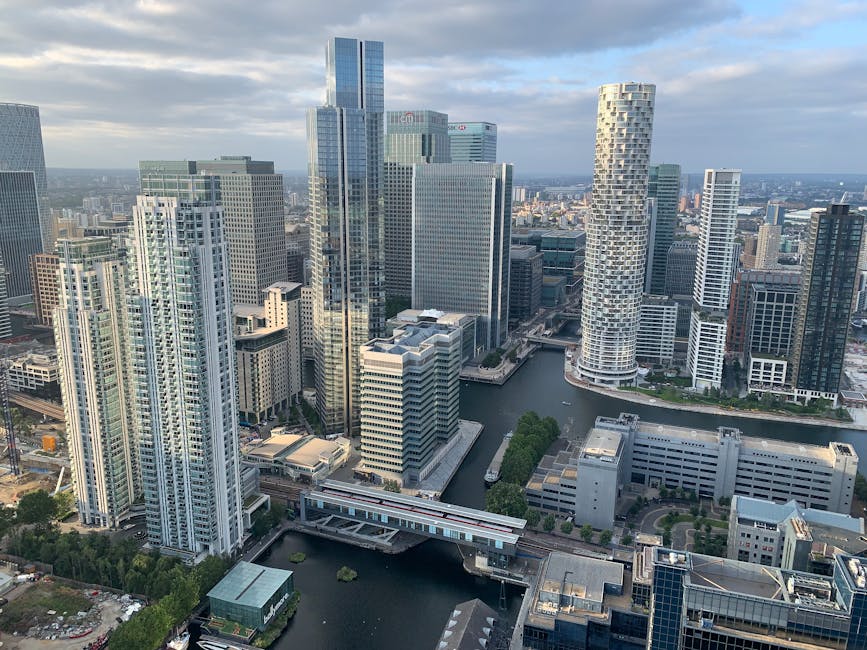 Aerial view of Canary Wharf in London showing multiple modern high-rise office buildings with glass facades surrounding a central waterway. The buildings include a mix of rectangular and uniquely shap