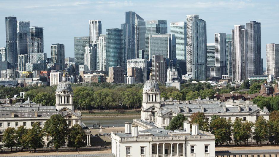 Aerial view of Canary Wharf in London showing multiple modern high-rise office buildings with glass facades surrounding a central waterway. The buildings include a mix of rectangular and uniquely shap