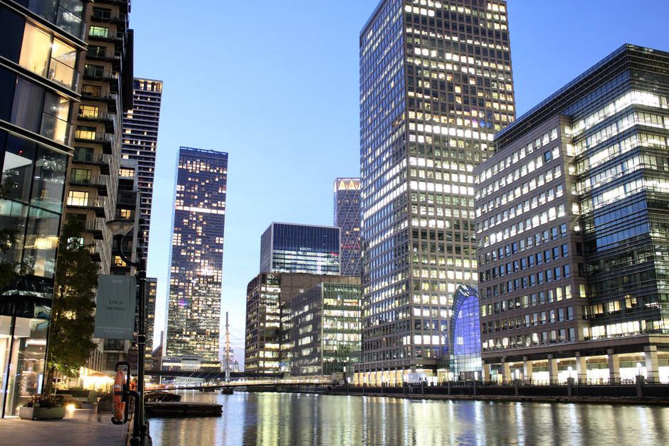 A wide view of the Canary Wharf financial district in London during early evening, featuring tall glass office buildings with illuminated windows reflecting on the waterway in the foreground. To the left, a modern building with large glass panels and a lit entrance is visible, while on the right, multiple skyscrapers with grid-like window patterns rise above. The sky is clear with a soft blue hue, and a few streetlights and potted plants along the pavement are visible. This setting exemplifies the urban environment where home relocation or furniture transport might involve navigating narrow access points and tight spaces near high-rise buildings, highlighting the importance of careful planning during removals services, as offered by Canary Wharf Removals.
