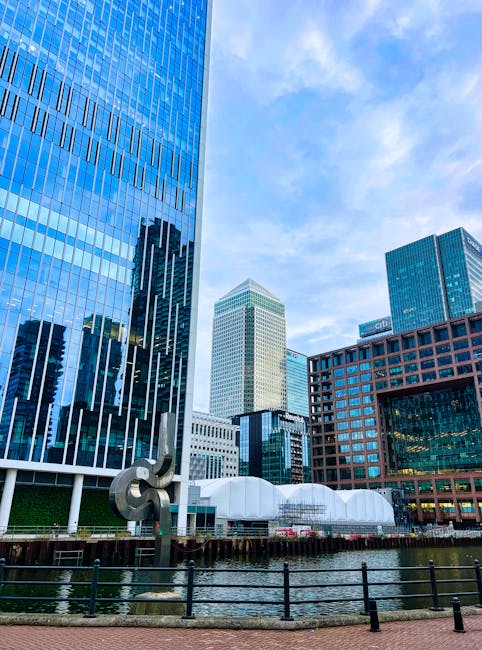 Photograph of a modern urban waterfront scene in Canary Wharf featuring several high-rise office buildings with reflective glass facades, including a tall blue-tinted tower and a beige structure with a grid-like window pattern, set against a partly cloudy sky. In the foreground, there is a black metal railing along the water's edge, with a small sculpture of intertwined metallic rings positioned near the railing. Behind the railing, a white canopy structure with rounded peaks is visible, and nearby, there is a loading area with a van being loaded with cardboard boxes and wrapped furniture, indicating a home relocation or furniture transport operation. The scene is well-lit with natural daylight, highlighting the busy moving process associated with professional removals in a city centre environment. This image reflects the logistical aspects of house removals in a commercial district, consistent with [PAGE_TITLE], provided by Canary Wharf Removals.