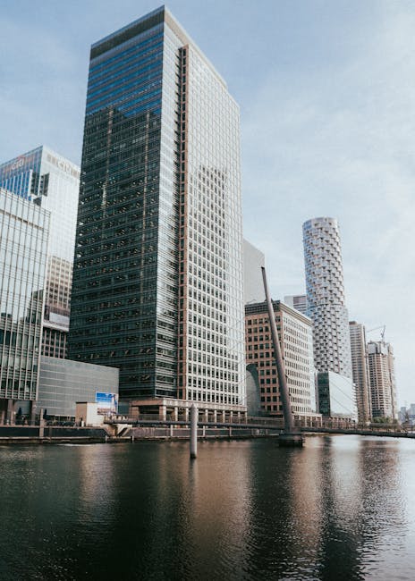 A view of tall modern office buildings along the Canary Wharf waterfront, with glass facades reflecting the sky, and construction cranes visible on some structures during daylight hours. In the foreground, the river waters are calm, with a low wall or quay separating the water from the buildings. The scene showcases urban architecture typical of a city centre with prominent commercial skyscrapers, which are relevant to house removals and relocation services. This setting is part of a moving guide for One Canada Square residents, illustrating the type of environment where furniture transport and packing and moving activities often take place. Canary Wharf Removals might be involved in the logistics of home relocation for clients working or living within these office towers, emphasizing the importance of careful loading and transport of belongings during house removals in the Canary Wharf area.