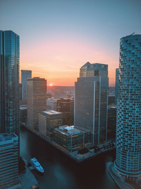 A high-angle view of Canary Wharf during sunset, showcasing a cluster of modern glass and steel skyscrapers with reflective surfaces. The river Thames is visible at the base of the buildings, with a white yacht docked along the quay. The sky is transitioning from warm orange and pink hues near the horizon to soft blue at the top, indicating the nearing end of the day. The buildings are part of a dense commercial area, and the scene captures the vibrant cityscape typical of London’s financial district. This image appears relevant for house removals or moving services in Canary Wharf, illustrating the urban environment where professionals like Canary Wharf Removals assist with home relocation, furniture transport, and moving logistics in this prominent area.