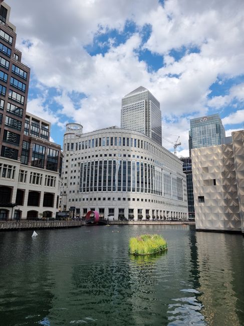 A view of tall modern office buildings along the Canary Wharf waterfront, with glass facades reflecting the sky, and construction cranes visible on some structures during daylight hours. In the foreground, the river waters are calm, with a low wall or quay separating the water from the buildings. The scene showcases urban architecture typical of a city centre with prominent commercial skyscrapers, which are relevant to house removals and relocation services. This setting is part of a moving guide for One Canada Square residents, illustrating the type of environment where furniture transport and packing and moving activities often take place. Canary Wharf Removals might be involved in the logistics of home relocation for clients working or living within these office towers, emphasizing the importance of careful loading and transport of belongings during house removals in the Canary Wharf area.